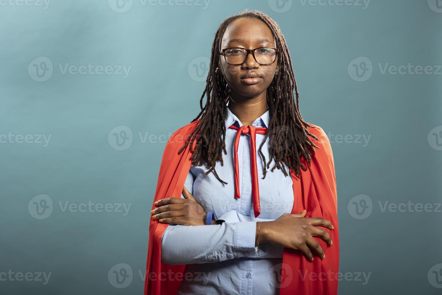 Portrait of female corporate superhero posing with her arms crossed and looking at camera. African american woman standing in blue background studio, expressing confidence and empowerment. photo