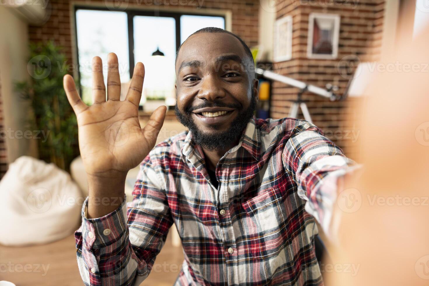 Happy bearded black man waves at camera, connecting remotely with colleague in modern apartment living room. African American freelancer doing greeting gesture during cheerful call from home. photo