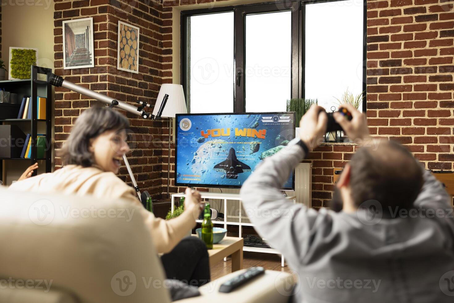 White man and woman relax together, celebrating shared game victory. Young caucsasian adults with raised hands, cheering and enjoying drinks on sofa while TV flashes winning message. photo