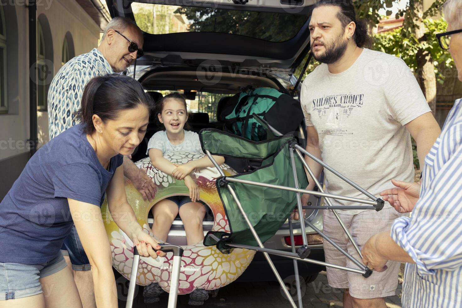 Grandparents help their son and daughter in law load the car trunk while a little girl watches with joy, big group excited about family getaway to the seaside. Relaxing road trip lifestyle. photo