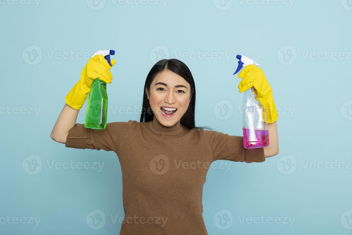 Energetic asian housekeeper in rubber gloves holding colorful cleaning sprays and flexing arms, promoting cleanliness with big smile. Enthusiastic female cleaner with pink and green liquid solutions. photo