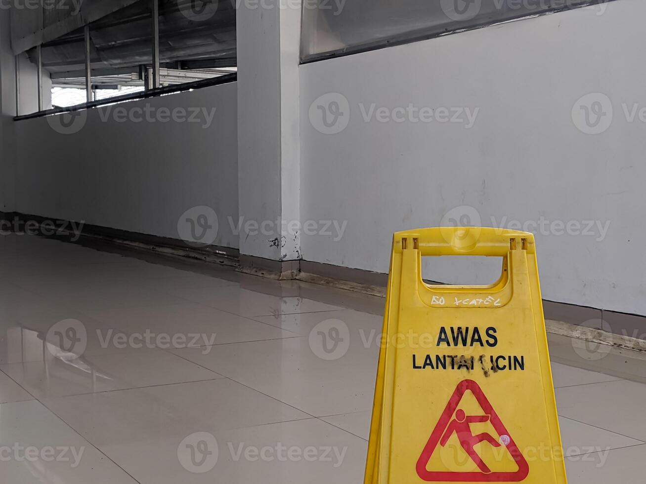 Yellow safety sign in Indonesian AWAS LANTAI LICIN, Caution Wet Floor, with a red falling figure symbol, indicating a slip hazard on a wet tiled floor in Banjarsari, West Java, Indonesia. photo