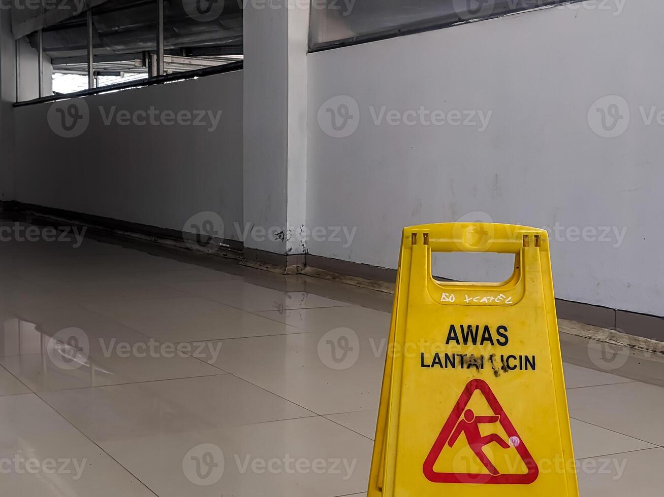 Bright yellow safety sign displaying AWAS LANTAI LICIN and a falling person icon, positioned on a reflective tiled floor, alerting to a wet and hazardous surface in Banjarsari, West Java. photo