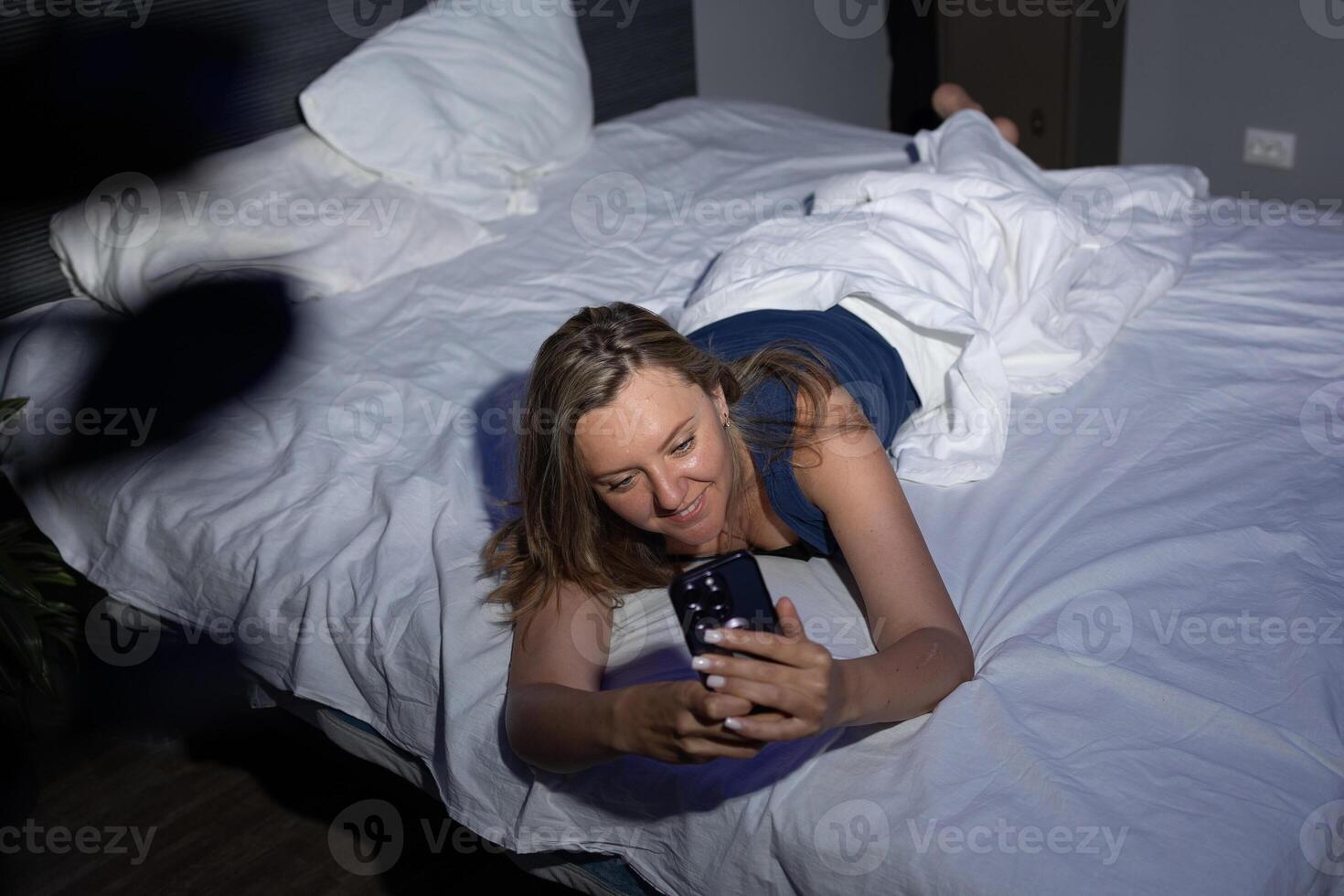 Smiling Woman Enjoys Late-Night Relaxation While Using Smartphone in Cozy Bedroom Setting photo