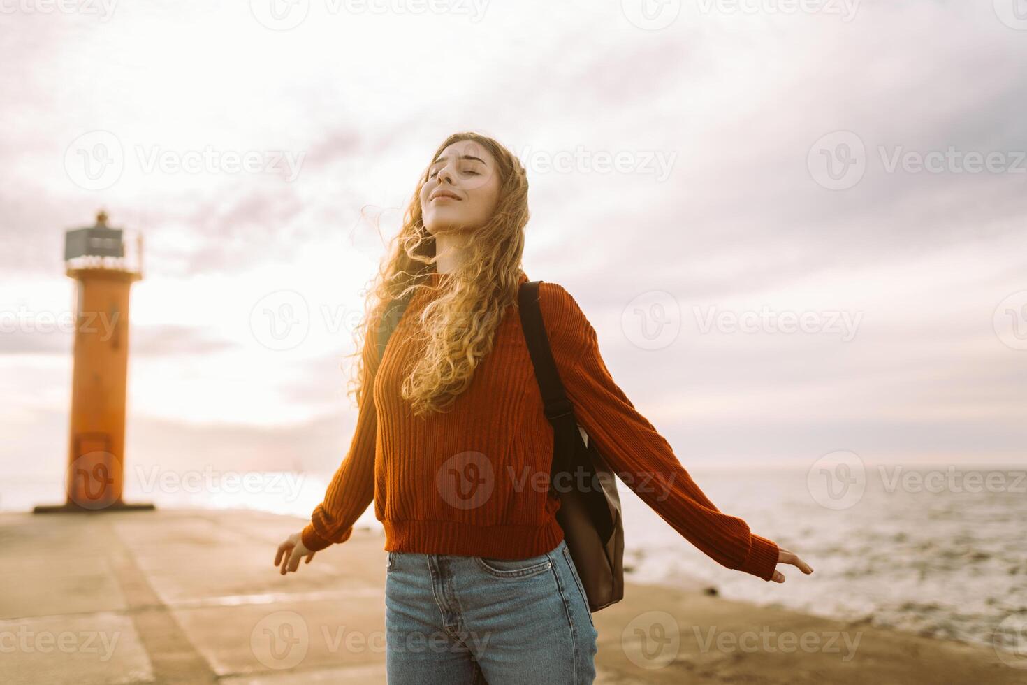 Woman enjoying breezy day near vibrant red lighthouse at sunset. Travel, weekend, lifestyle photo