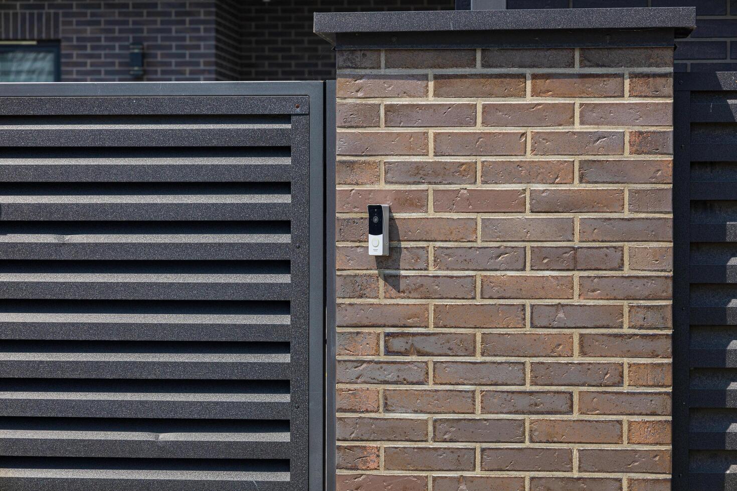 A contemporary intercom is mounted on a brick wall adjacent to a slatted gate in a well-maintained suburban area photo