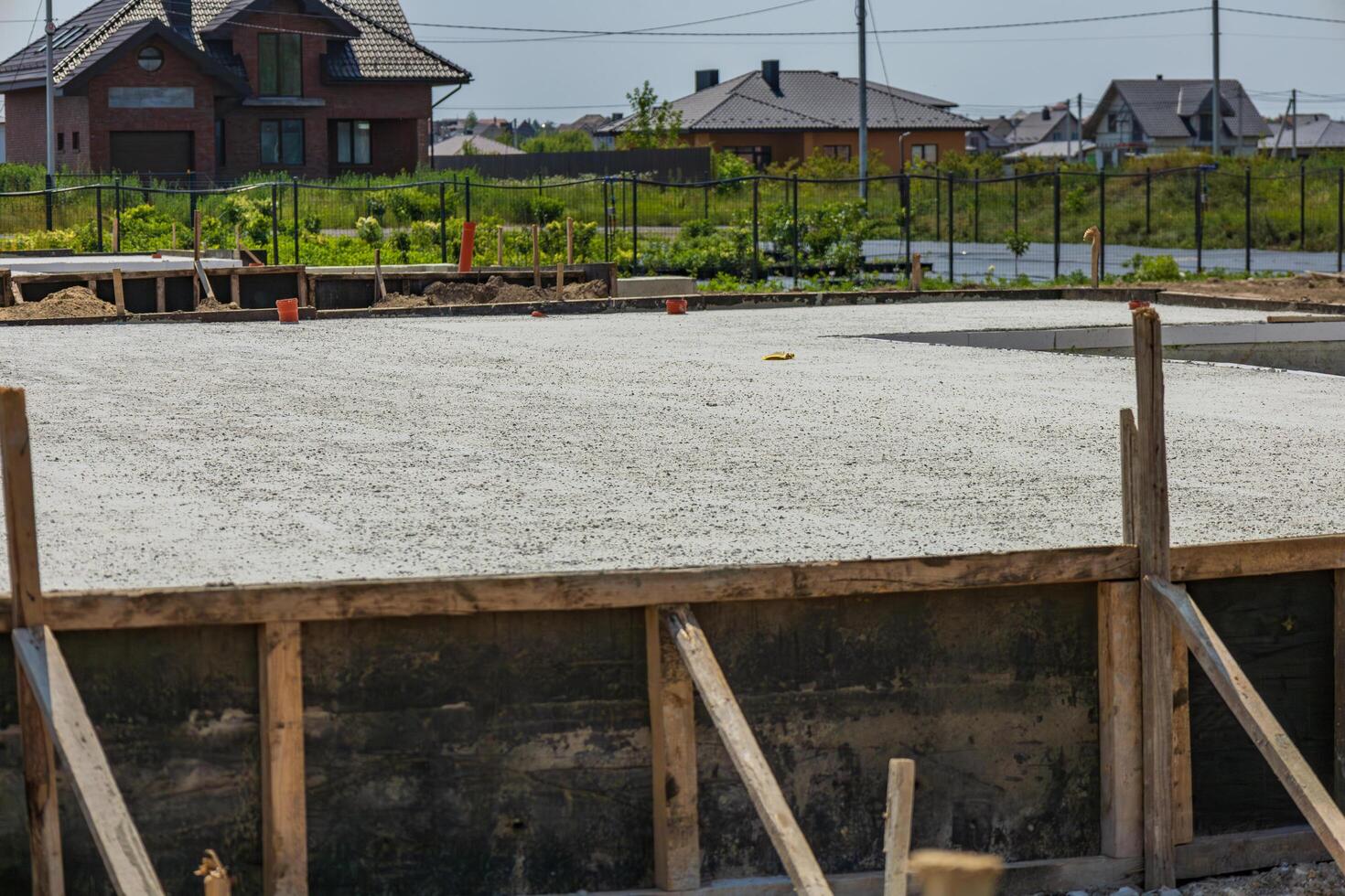 A concrete foundation has been freshly poured at a construction site surrounded by houses and greenery under a bright sky photo