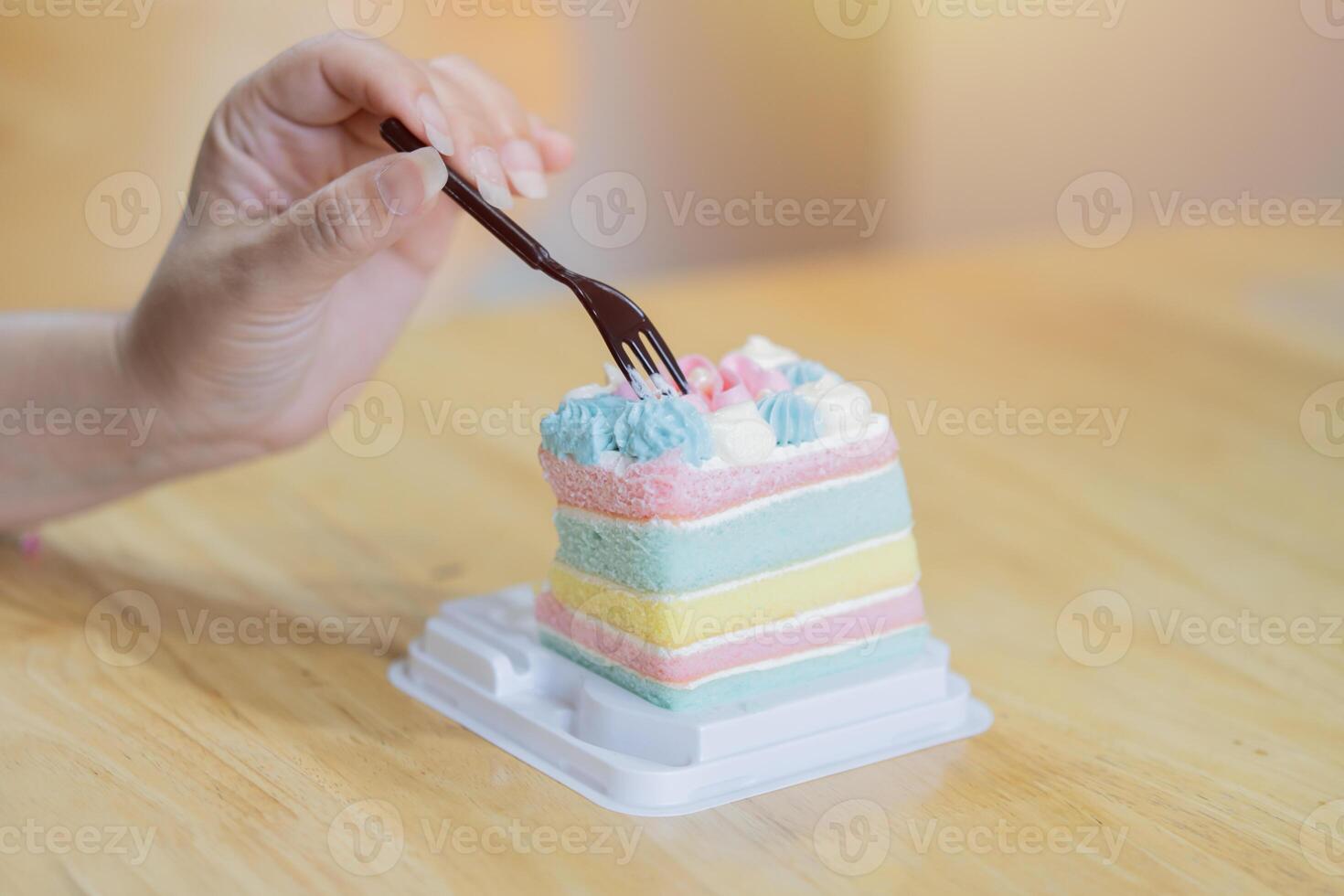 Close-up image of a woman hand eating a painbow cake in plastic tray over wooden table at cafe. photo