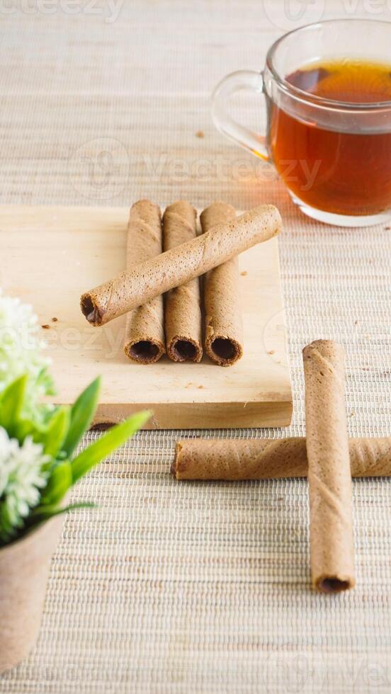 Crispy chocolate wafer roll on a wooden board with a cup of tea for enjoyment isolated on wooden textured background photo