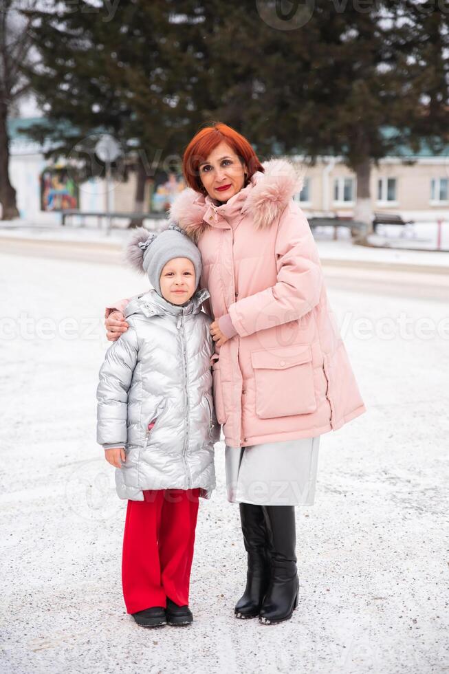 A Russian mother and daughter smiling together outdoors, dressed in matching coats. The daughter is standing next to her mother, both happy and enjoying a sunny day photo