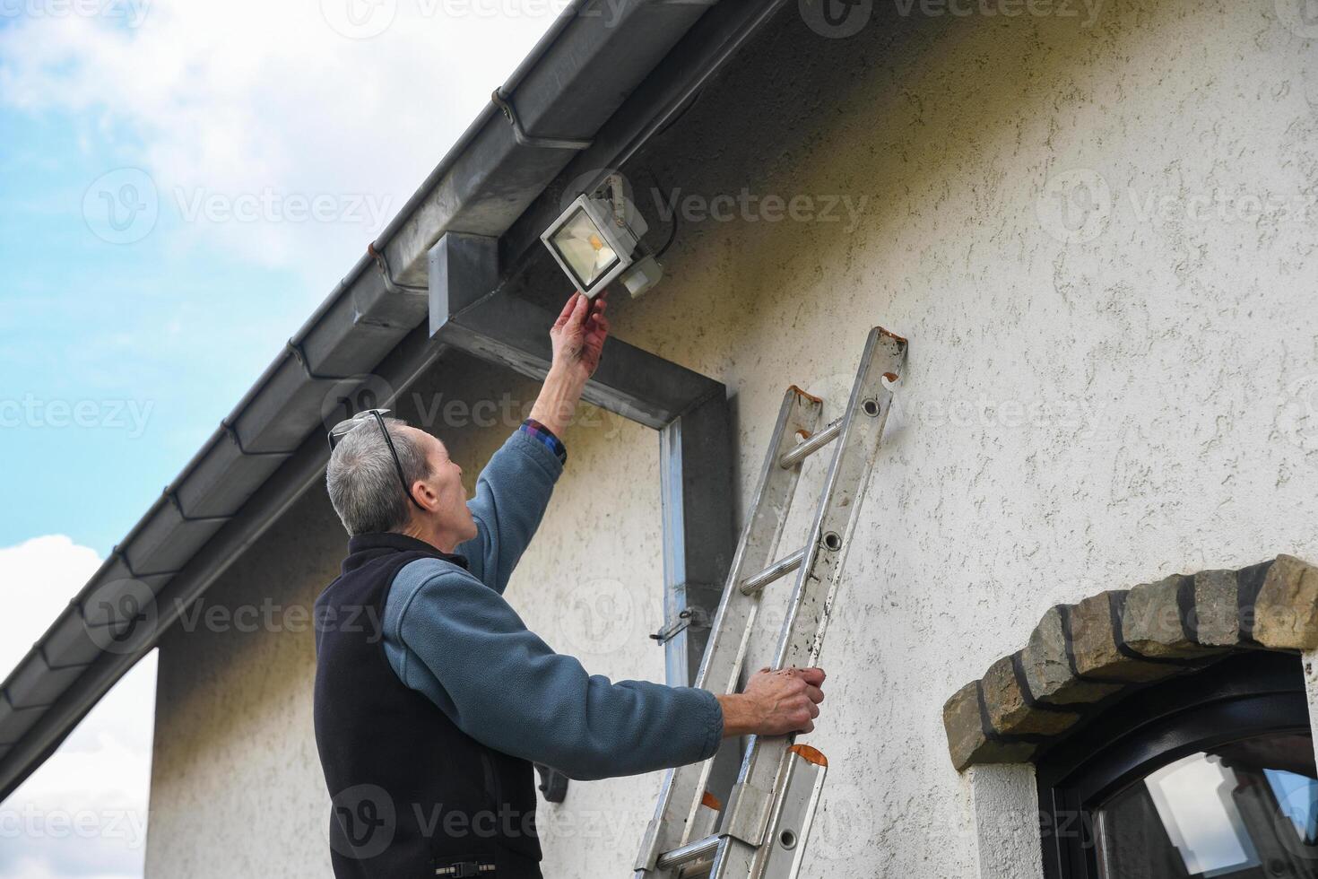 Electrician reaches for motion sensor LED floodlight while standing on ladder installing it on facade of house under gutter to improve outdoor lighting photo