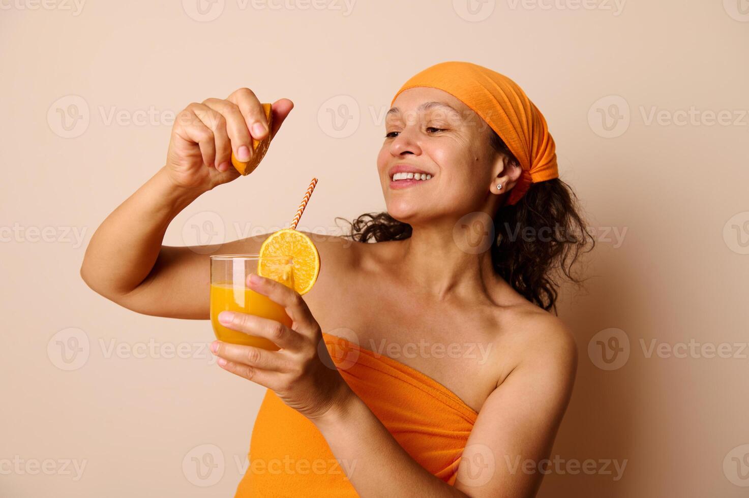 Smiling Woman Pouring Orange Juice While Holding a Glass, Dressed in Orange Against a Beige Background photo