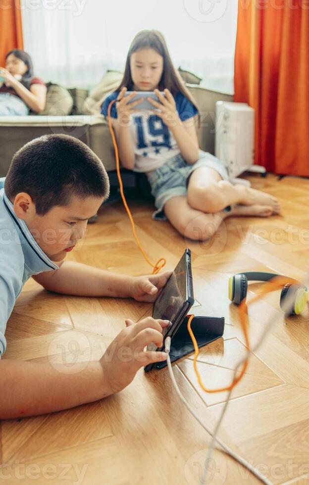 boy lying on wooden floor playing with tablet while two girls relax on sofa using smartphones with orange curtains and daylight, technology education leisure, vertical photo