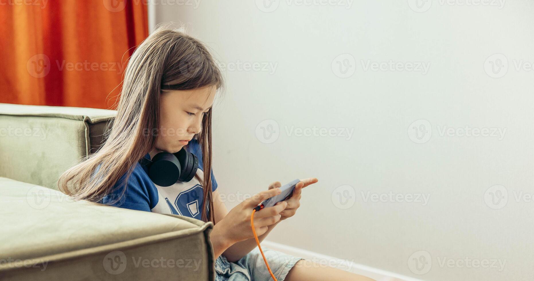 Asian half-breed girl near green sofa using smartphone, headphones, home interior with soft light, technology gadgets youth lifestyle, copy space photo
