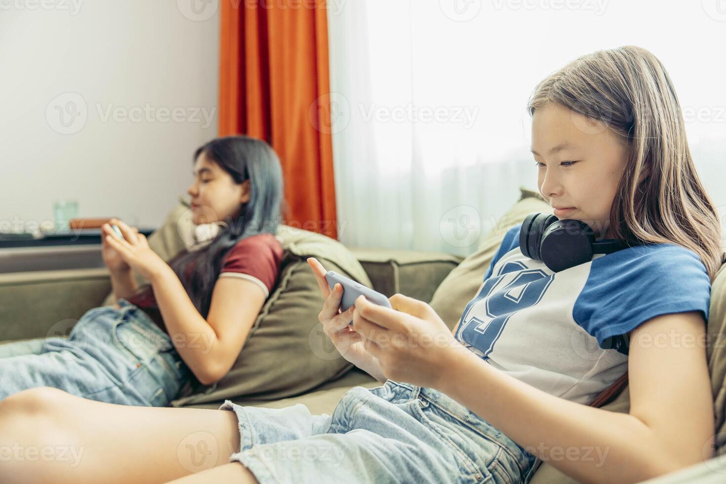 two girls relaxing on sofa using smartphone, headphones, casual home setting bright curtains and natural light window modern lifestyle, technology education entertainment photo