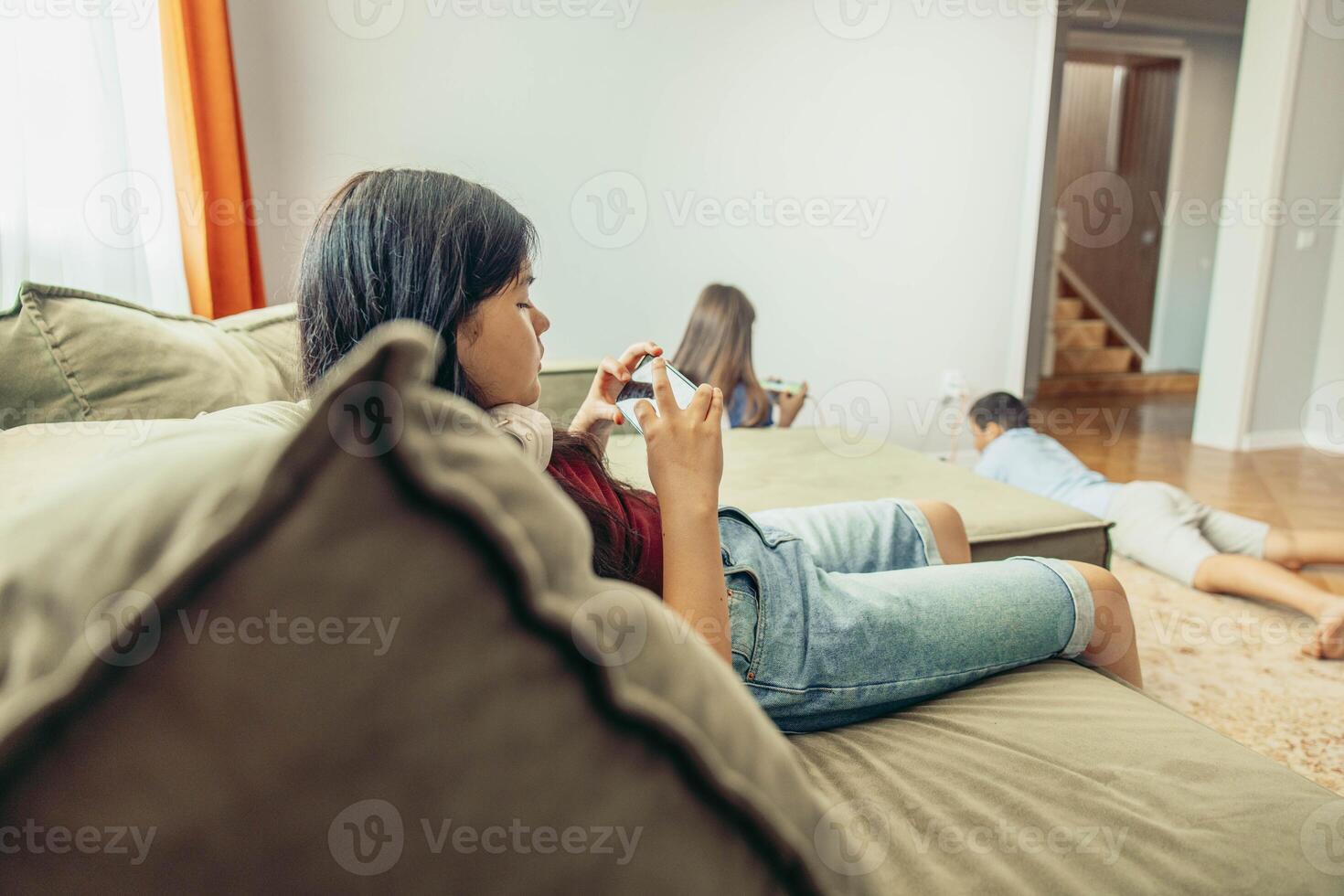 Asian teen girl with smartphone, boy and girl on wooden floor playing with tablet with orange curtains, technology education leisure, power outlet with charging cables photo