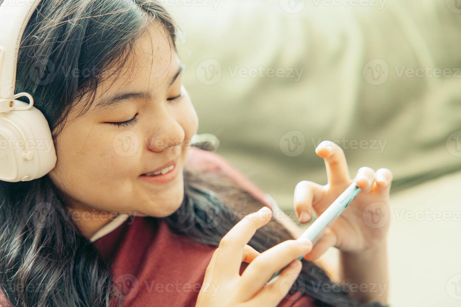 close up of smiling Asian girl lying on green sofa using smartphone with headphones around neck focused expression home interior,soft light, technology gadgets, lifestyle photo