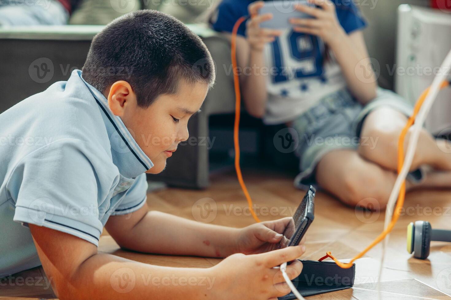 boy lying on wooden floor playing with tablet while Unrecognizable girl relax near sofa using smartphones with orange curtains and daylight, technology education, game photo