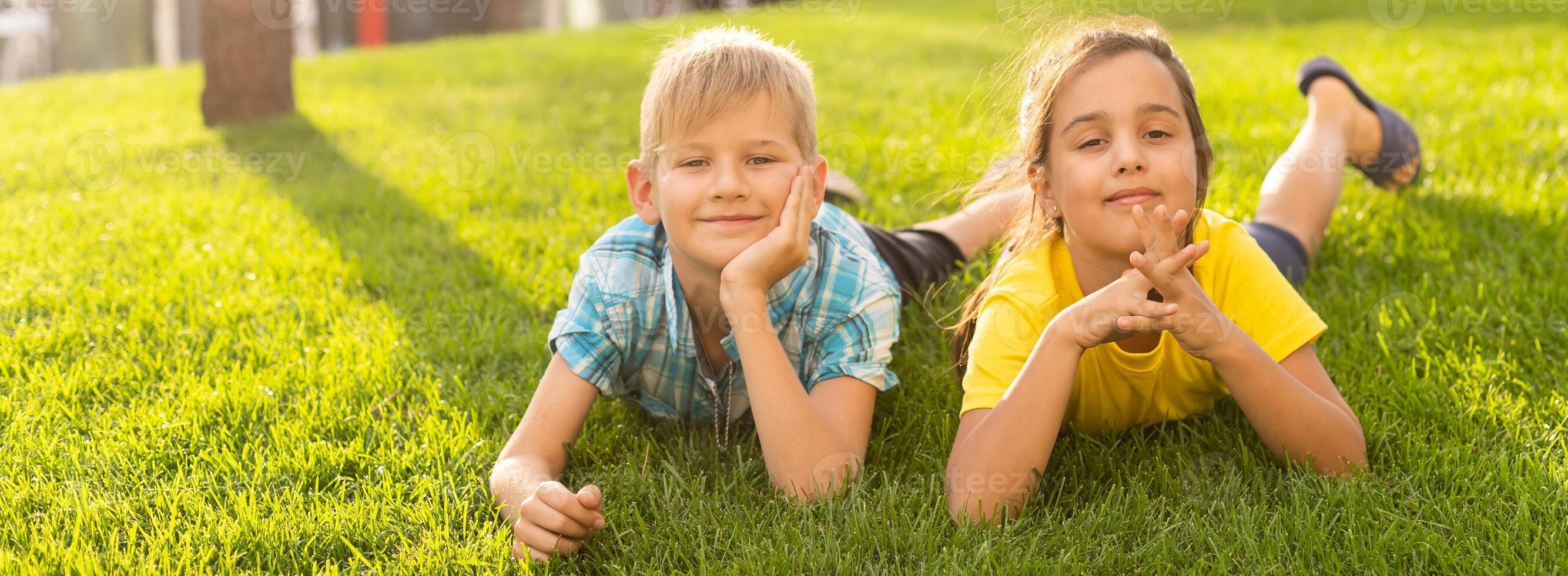 Happy peaceful childhood. Two happy little children - a boy and a girl in summer clothes are lying on a green lawn in a summer park. Summer kids fashion. Summer holidays, vacation. photo