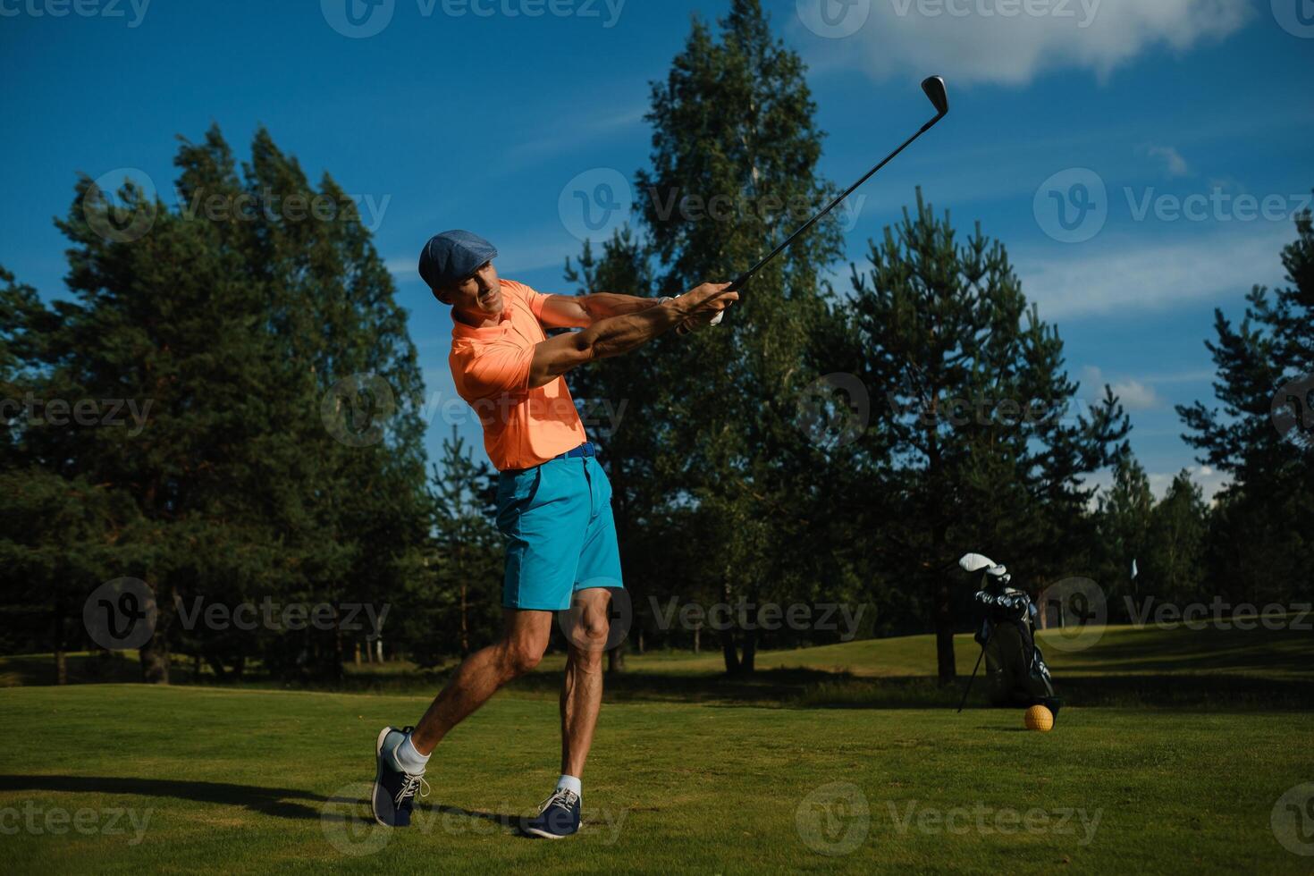 Golfer swings club on a sunny day at a lush green course surrounded by tall trees and blue skies photo