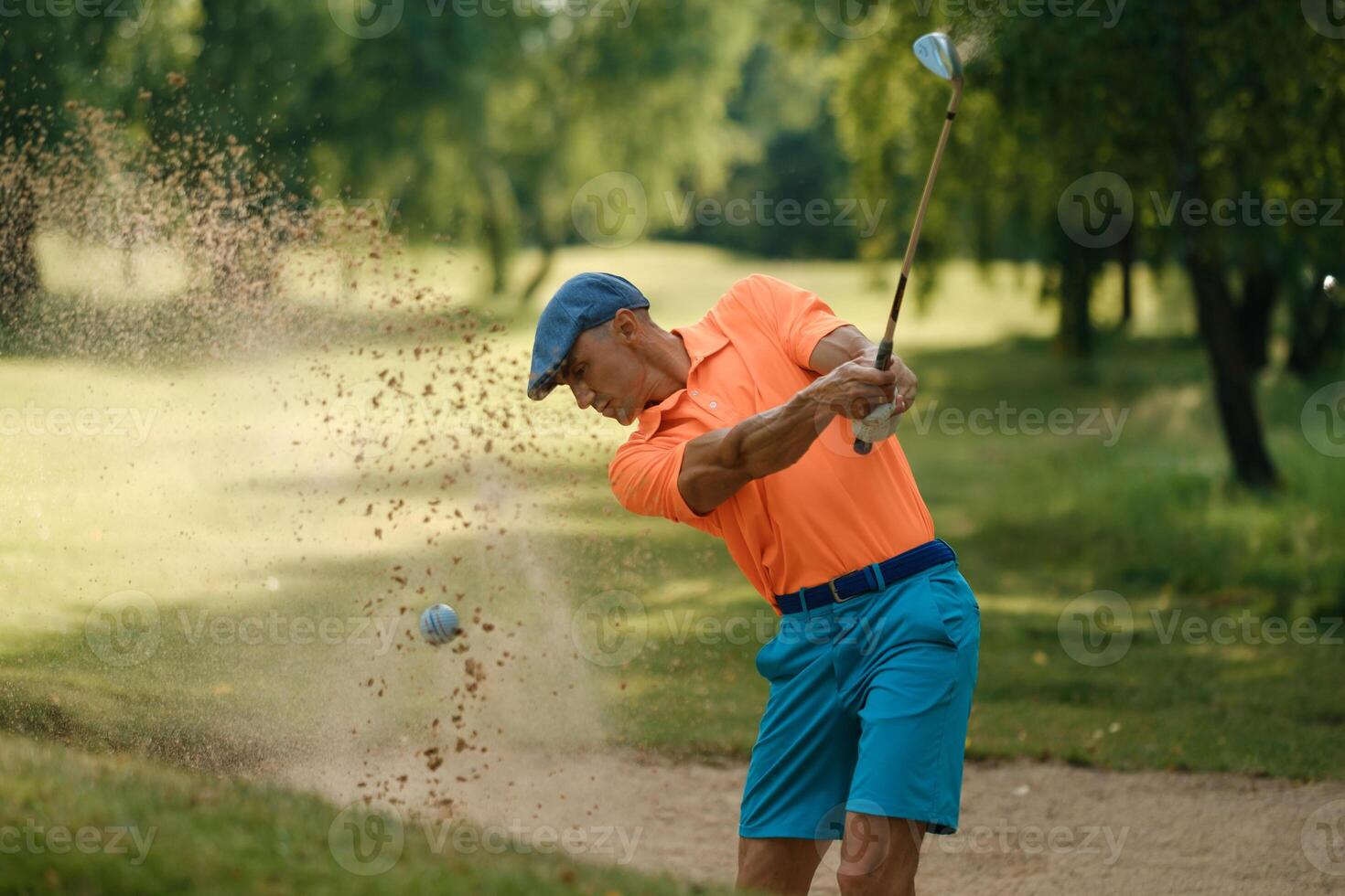 Golf player makes a powerful swing from a sand trap on a sunny day at a lush green golf course surrounded by trees photo