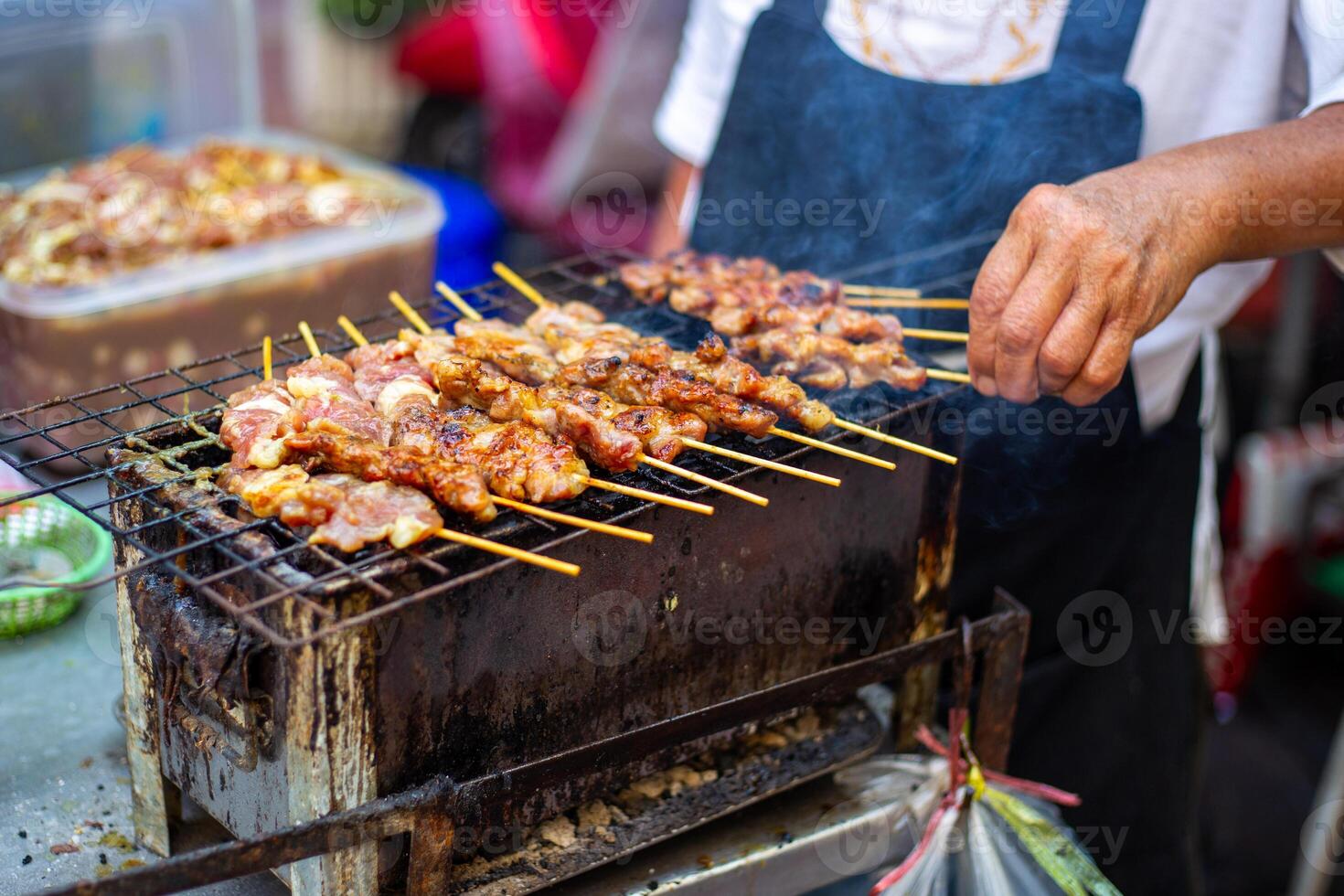 Close-up of a street food vendor grilling marinated meat skewers on a charcoal grill, a popular sight at Asian street markets photo