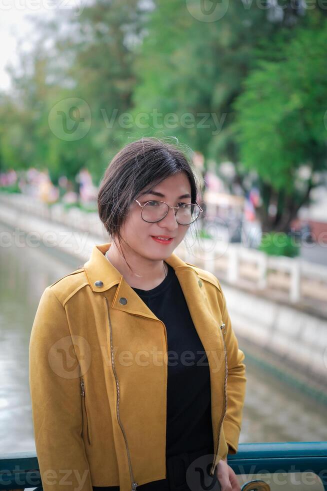 A woman wearing glasses and a yellow jacket standing near a river photo