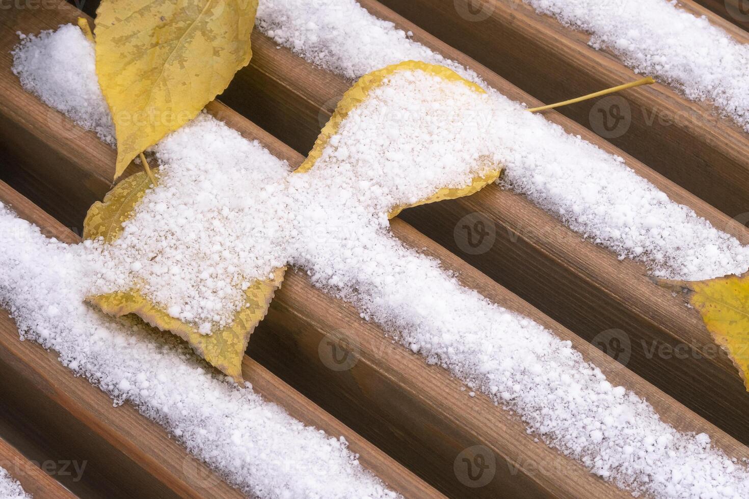 Wooden bench seat closeup. Covered with snow and yellow autumn leaves. Selective focus. photo