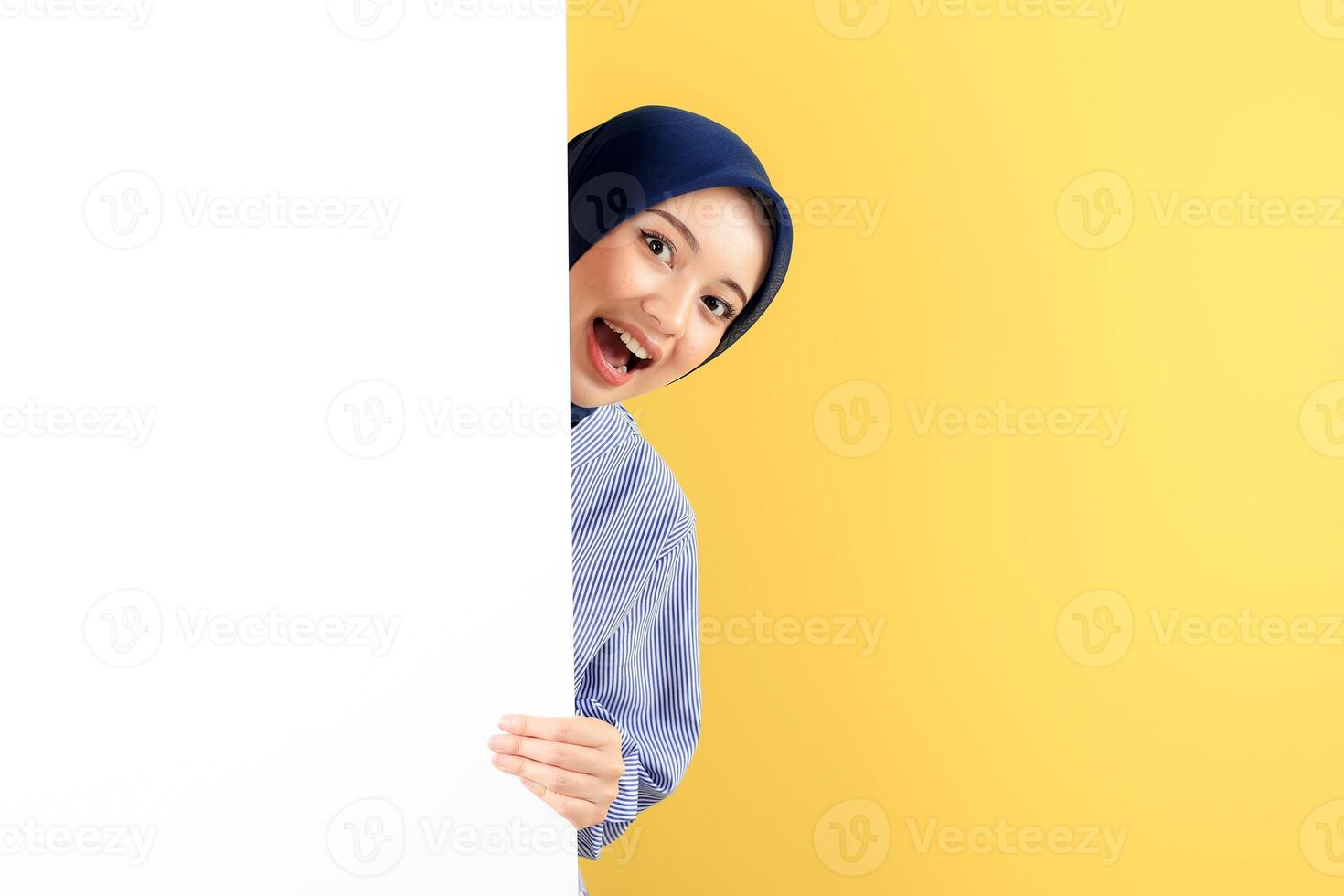 Happy Smiling Young Asian Muslim Woman with Hijab Peeking Out Behind a White Wall photo