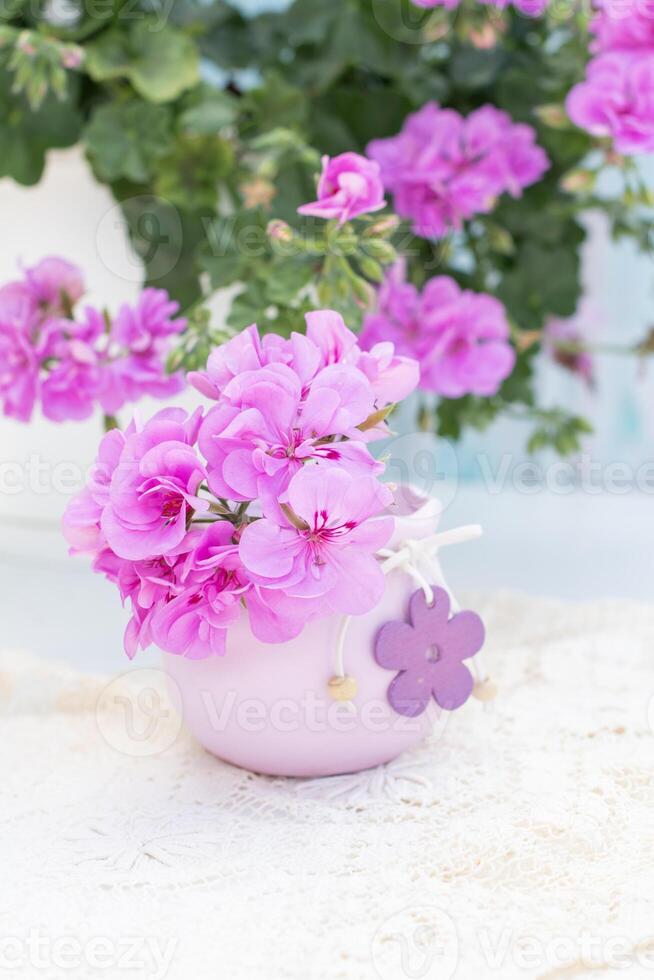 Pink geranium flowers arranged in a decorative purple vase with wooden flower embellishments, placed on a white doily, with additional geraniums visible in soft focus in the background,vertical photo