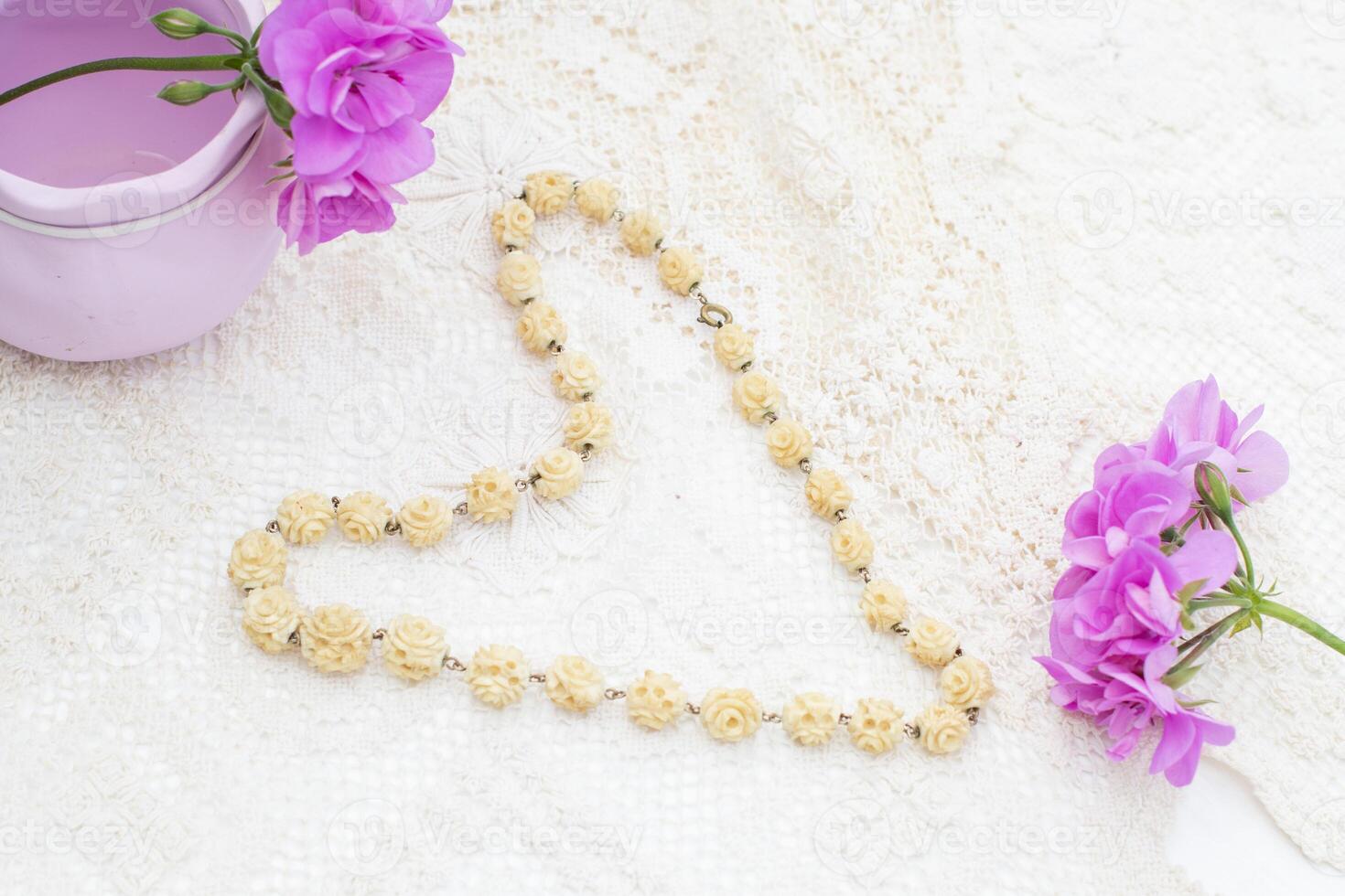 Vintage ivory necklace with rose-shaped beads lies on a white lace tablecloth in the shape of a heart, next to purple geranium flowers and a lilac vase, creating a delicate and romantic composition, photo