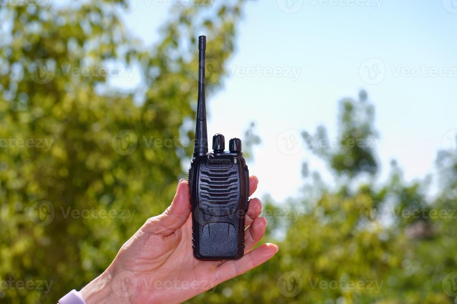 Handheld walkie talkie being held against a sunny outdoor background with greenery photo