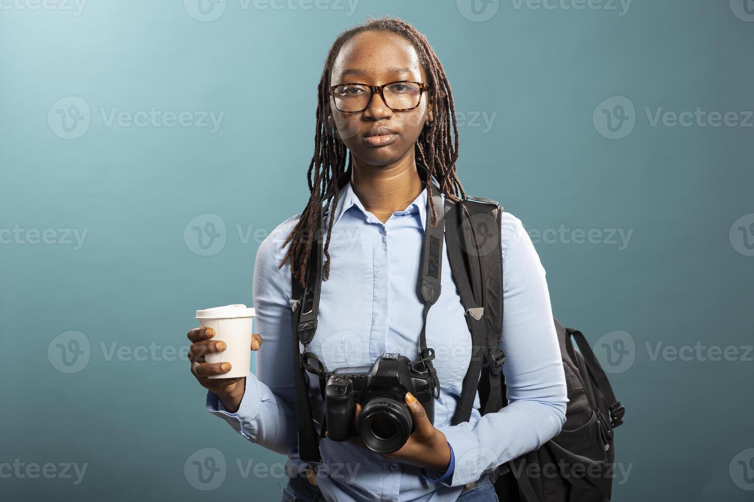 Young woman poses in studio, dressed casually with dslr camera hanging on her neck and coffee in hand. African american female model shows a focused yet professional photographer look. photo