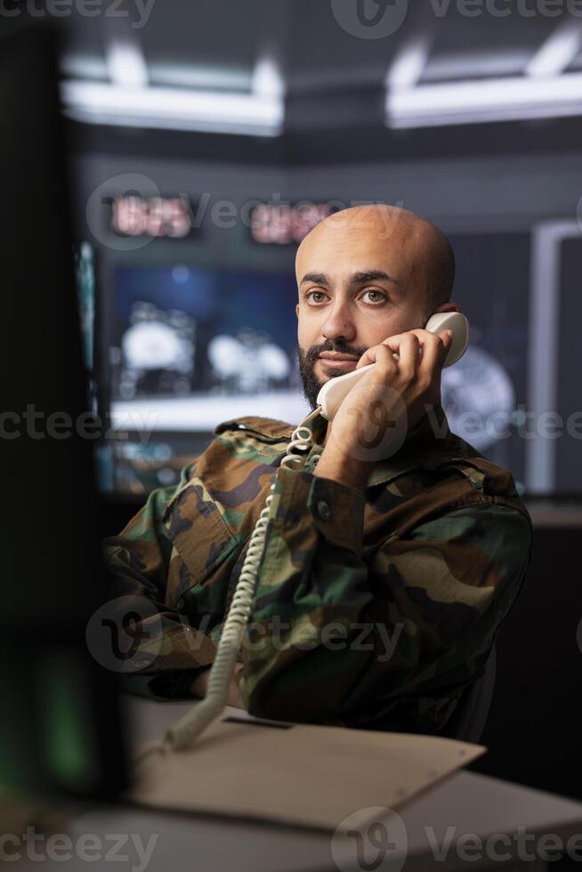 Military officer looking at surveillance footage, analyzing data to allocate resources to units based on mission needs, making telephone call. Agent examining data on screen and talking on phone photo