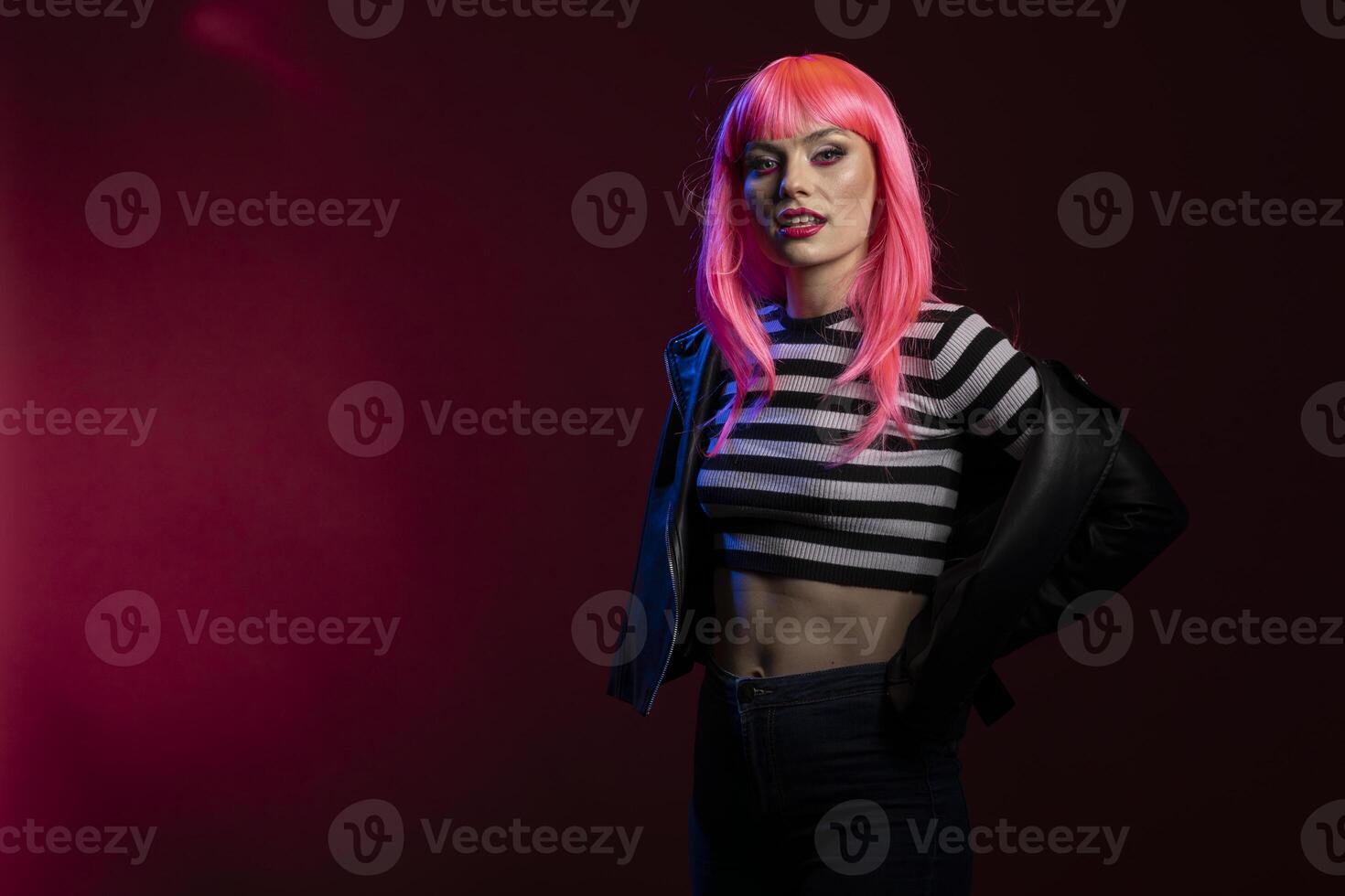 Stylish beautiful woman with vibrant pink hair and striped crop top posing in dark studio with neon lights. Attractive female model standing with attitude in bold fashion statement outfit. photo