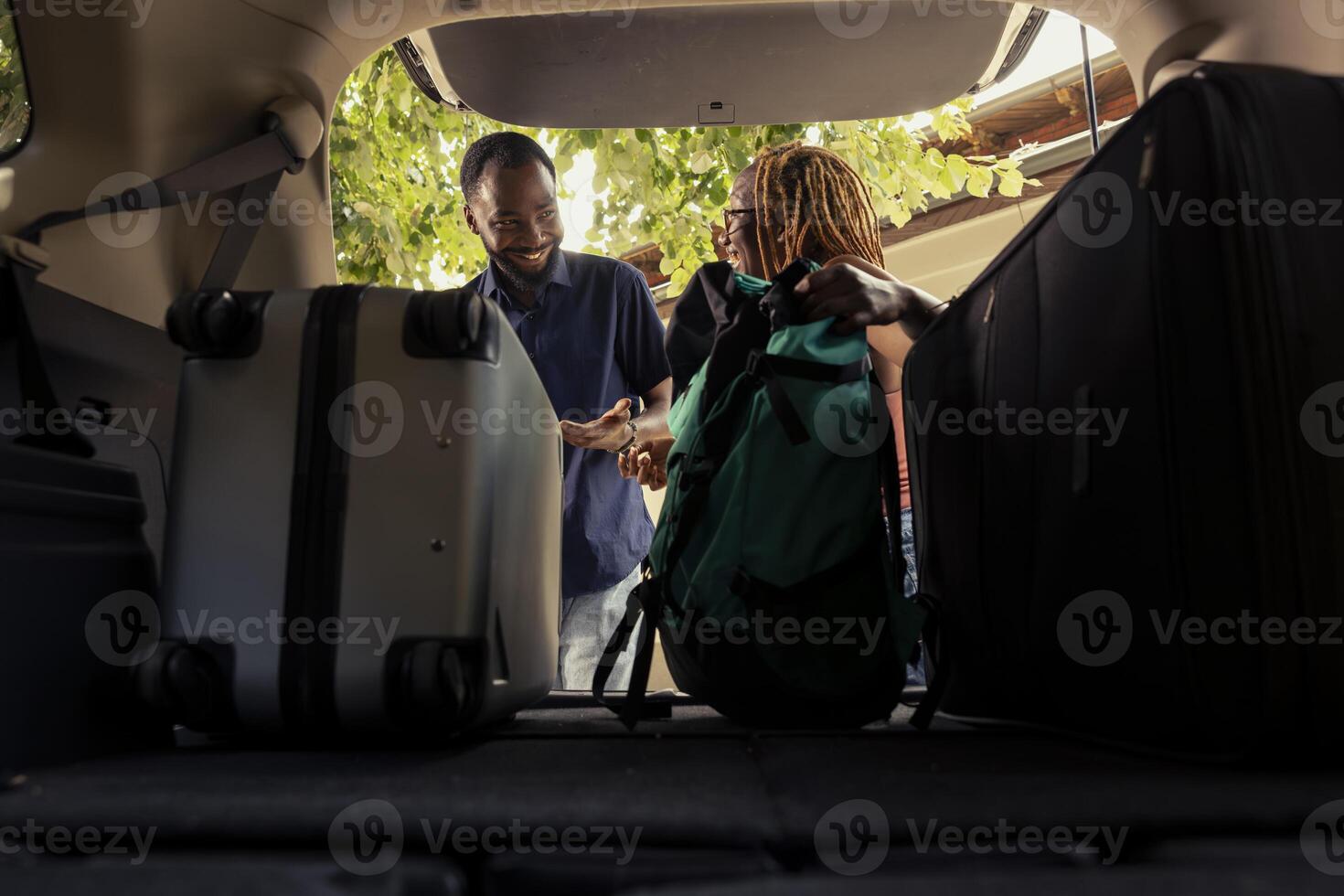 Black happy couple standing by their packed car and loading the trunk with suitcase under warm summer light. Happy life partners starting their relaxing road trip adventure together. photo