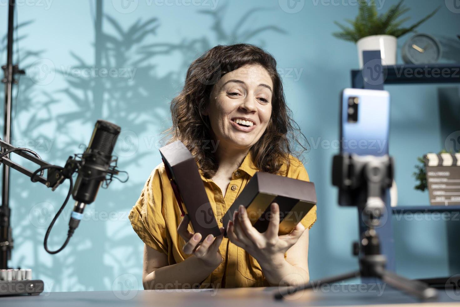 Social media influencer filming giveaway for audience using cellphone on tripod, holding gift box, explaining how to win prize. Jolly content creator running contest, filming with mobile phone photo