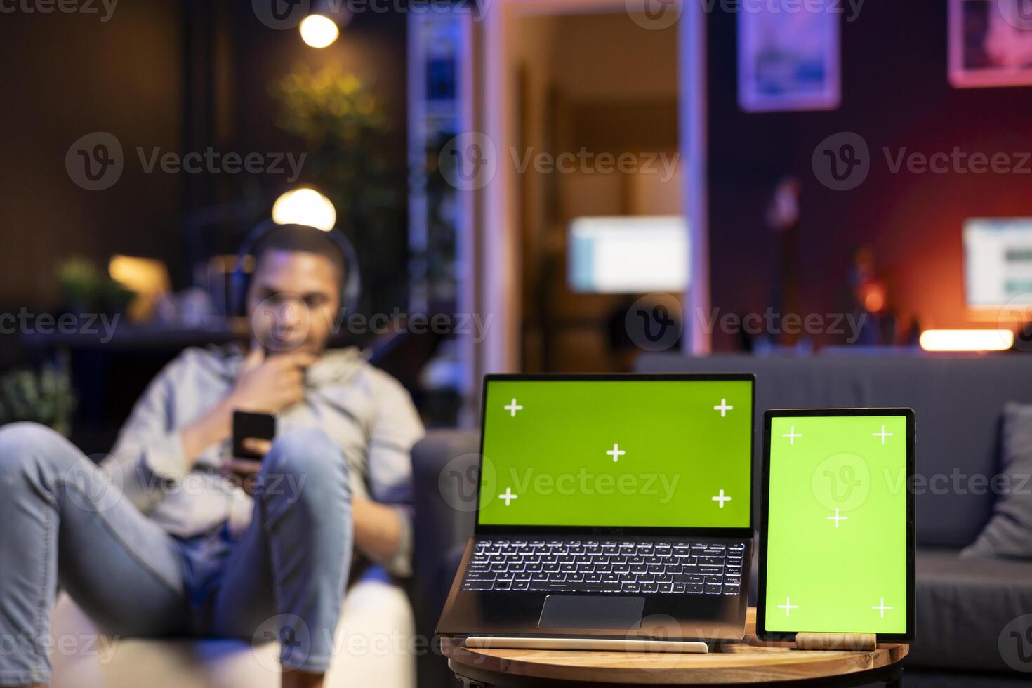 Young relaxed man with headset having a green screen in cozy living room, stylish space decorated with furniture and neon lights. Casual guy relaxing on a bean bag at home, multitasking. photo