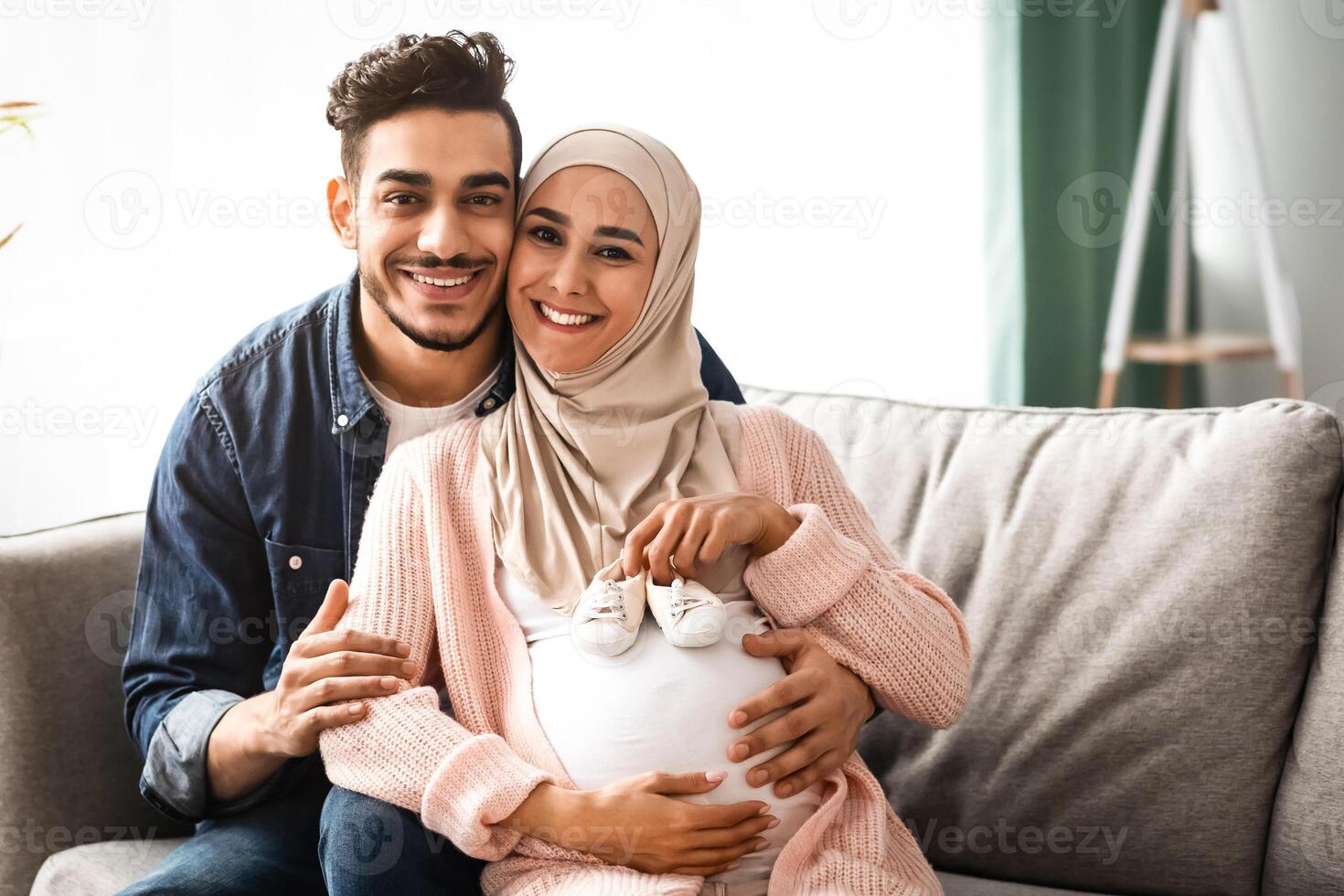 Young Muslim Family Expecting For Baby, Woman In Hijab Holding Small Baby Shoes Near Belly While Relaxing On Couch At Home With Her Husband, Islamic Spouses Enjoying Future Parenthood, Copy Space photo