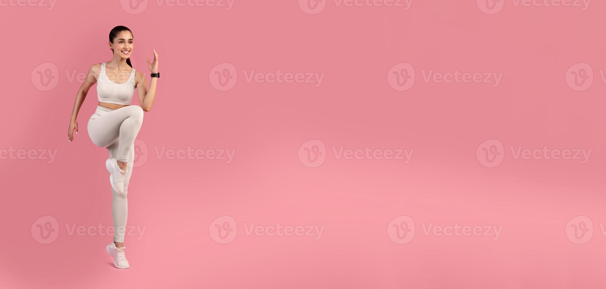 Aerobics Sport Exercises And Healthy Lifestyle. Portrait Of Smiling Slim Young Lady Lifting Leg Up, Doing High Knees Cardio Exercise Standing Isolated On Pink Studio Background, Full Length photo