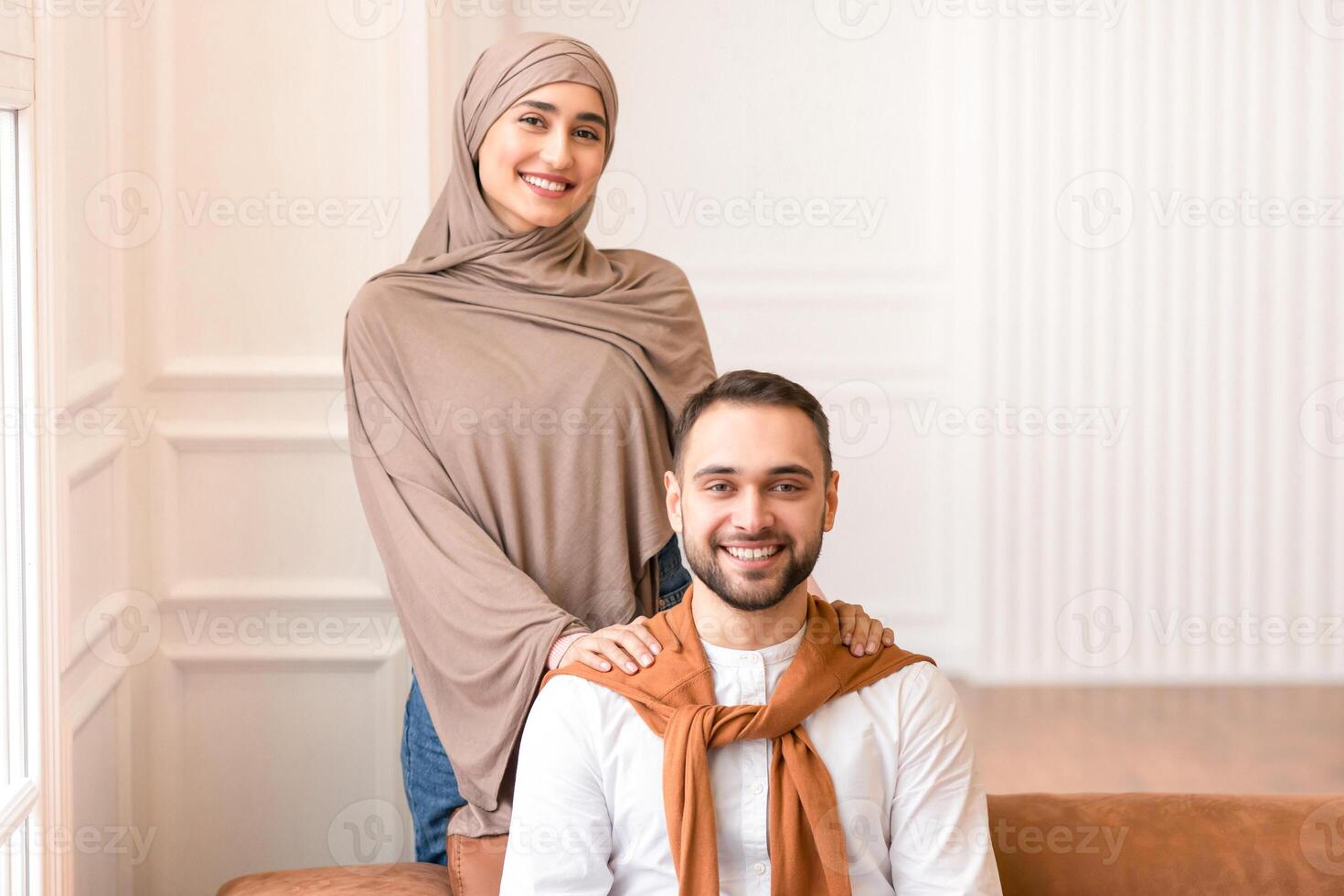 Married Middle Eastern Couple Posing Together Smiling To Camera At Home. Gladful Muslim Wife Wearing Hijab Hugging Her Husband Indoors. Modern Arabic Family Portrait Concept photo
