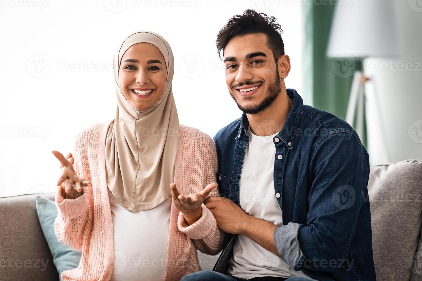 Happy News. Cheerful Pregnant Muslim Couple Talking At Camera, Making Pregnancy Or Baby Gender Announcement While Sitting On Couch At Home, Happy Islamic Spouses Smiling And Enjoying Future Parenthood photo