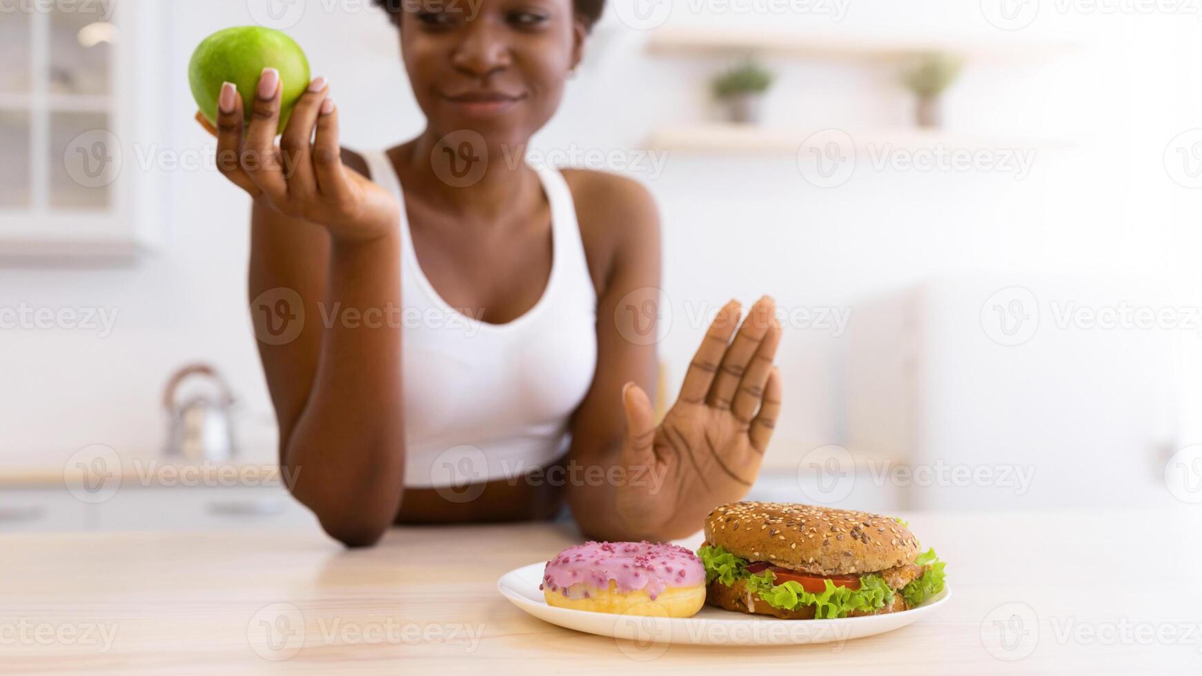 Black Woman Refusing To Eat Unhealthy Burger And Donut Choosing Healthy Apple Fruit Sitting In Modern Kitchen At Home. No Cheat Meal, Stop Eating Junk Food Concept. Selective Focus, Shallow Depth photo