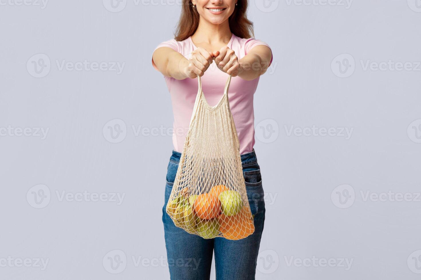 No Plastic For Shopping. Young Woman Holding Reusable Eco Mesh Bag With Fresh Organic Fruits, Cropped Image Of Smiling Lady Carrying Groceries, Standing Over Light Studio Background, Copy Space photo