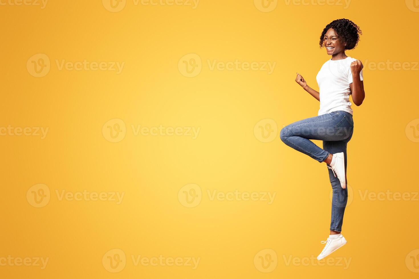 Cute black lady winner jumping up in the air and showing funny figures, having good day, showing positive emotions over orange background. African american woman posing at studio, having fun photo