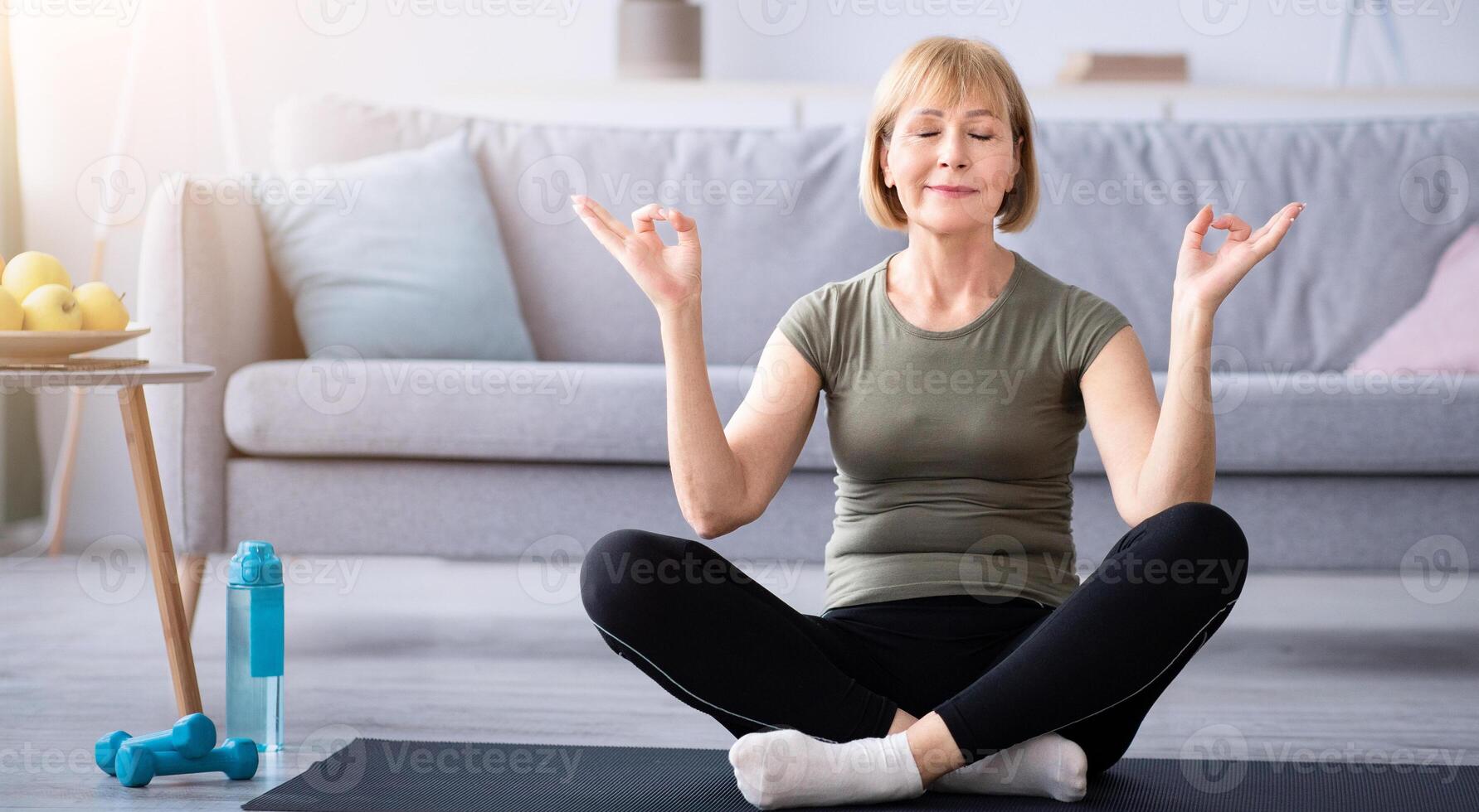Full length of mature woman meditating with closed eyes, making gyan mudra with her hands at home. Peaceful senior lady doing yoga, finding inner balance during covid lockdown photo