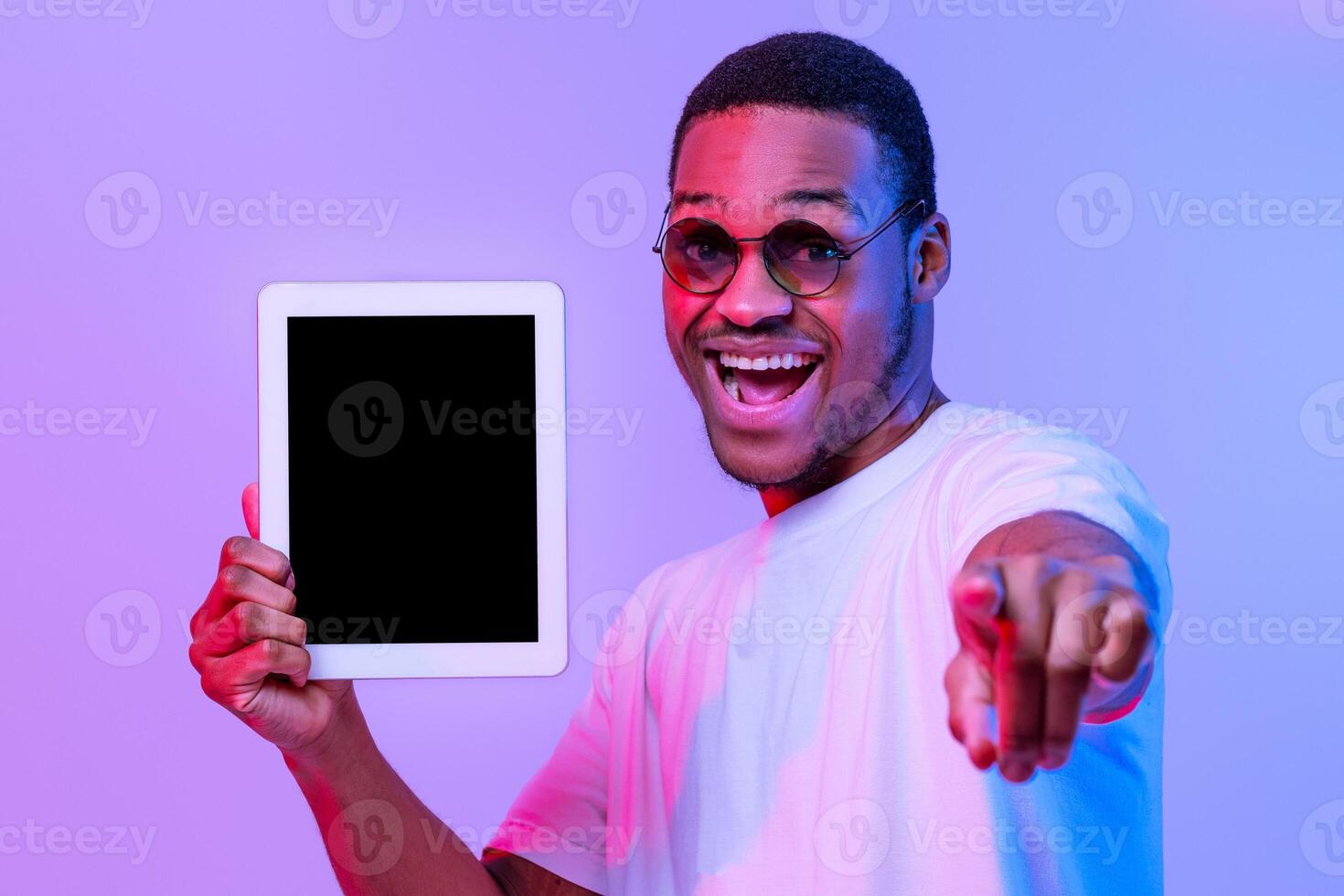 App For You. Joyful African American Guy Holding Digital Tablet With Black Screen And Pointing At Camera, Cheerful Millennial Man Standing Under Neon Light Over Purple Studio Background, Mockup Image photo
