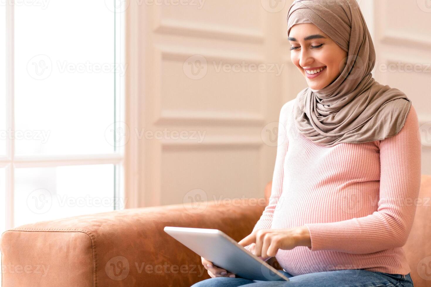 Happy Pregnant Muslim Woman Using Digital Tablet Reading Blog About Pregnancy Online Sitting On Sofa At Home. Middle Eastern Female In Hijab Browsing Internet. People And Technology Concept photo