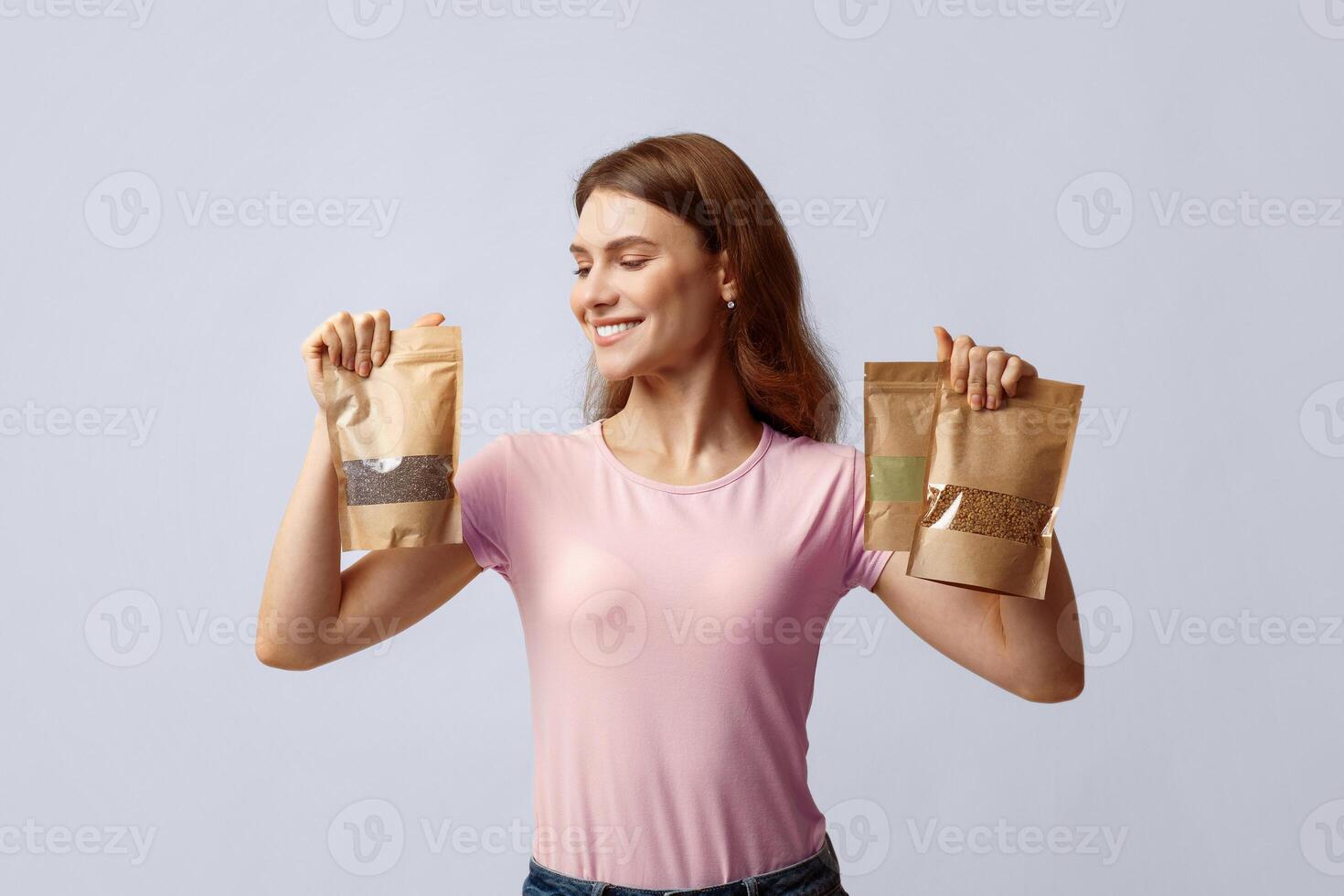 Happy young woman holding brown kraft paper doypack bags with weight groceries, enjoying eco-friendly shopping, smiling millennial lady standing over grey studio background, copy space photo