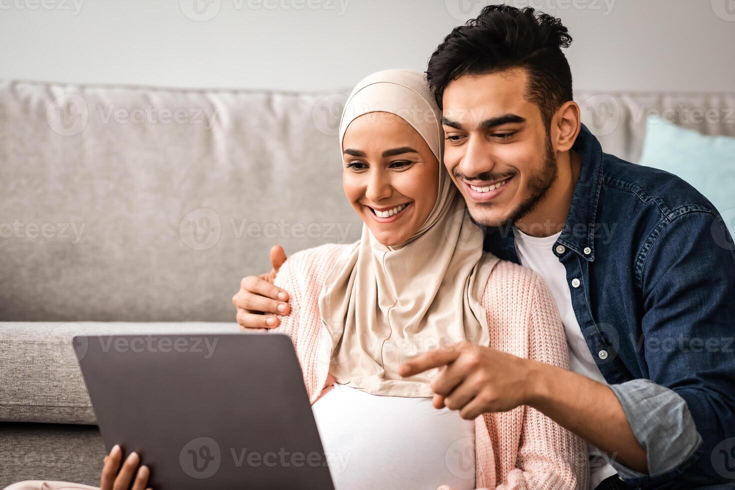 Cheerful Pregnant Muslim Woman In Hijab And Her Husband Using Laptop At Home Together, Middle Eastern Parents Shopping Online With Computer Or Watching Movie, Man Pointing At Screen, Closeup photo